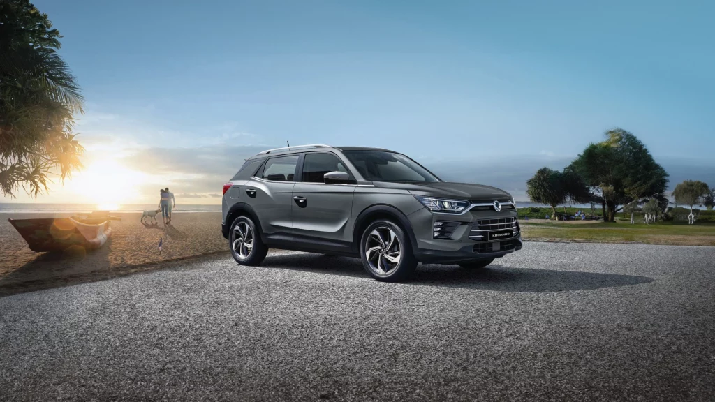 En KGM E-Korando SUV i grå farge parkert ved en strand i solnedgangen. Bilen har et moderne og elegant design med fremtredende frontlykter og felger. I bakgrunnen sees en strand med mennesker som går tur, samt noen trær og en liten båt.