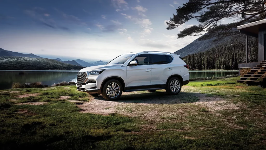 En hvit KGM Rexton SUV parkert ved et idyllisk innsjølandskap, omgitt av fjell og skog. Bilen har et moderne og robust design med en markant grill og stilige felger, og står plassert på en gresskledd bakke under en klar himmel.